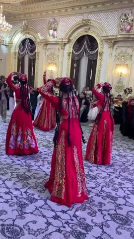 A group of women in traditional red dresses with gold embroidery are dancing in a large, ornate room. The room has high ceilings with intricate gold molding and chandeliers. Large curtains hang from the walls. The floor is covered in a patterned carpet. In the background, other people are seated at tables, watching the dancers. The dancers have their hair in long braids and are wearing matching headpieces. They are performing synchronized movements, with their arms raised and bodies in graceful poses. The lighting is bright and the atmosphere appears festive.