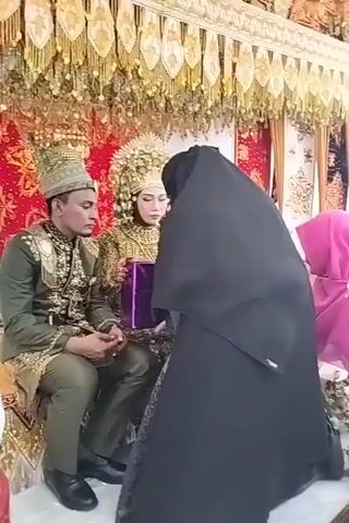 A wedding ceremony is taking place. A man and a woman, dressed in elaborate traditional wedding attire, are seated on a decorated platform. The man is wearing a green and gold embroidered suit with a matching hat. The woman is wearing a gold, intricately designed outfit with a tall, ornate headdress. Between them, they are holding a purple gift box. A woman in a black hijab is standing in front of them, and another woman wearing a pink hijab and a pink embroidered dress is partially visible to the right, leaning forward. The backdrop is adorned with golden fabric, red fabric with gold patterns, and white fabric with gold patterns.