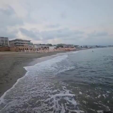 The video opens with a shot of a calm beach, the ocean gently lapping at the shore. The camera pans slowly to the right, revealing a rocky outcrop in the foreground. In the distance, a line of buildings and beach umbrellas can be seen along the coastline. The sky is overcast with a few scattered clouds. As the camera continues to pan, the focus shifts to the open sea, where several ships are visible on the horizon. The waves continue to break against the rocks, creating a soothing soundscape. The overall mood of the video is peaceful and serene, capturing the beauty of a quiet beach on a cloudy day.