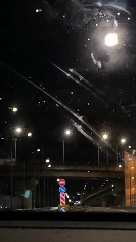 The video shows a night-time view from inside a vehicle, likely from the windshield. The scene is dominated by darkness, with streetlights creating bright circles of light. The road ahead is lined with more streetlights, creating a sense of perspective. There are traffic signs visible, including a red circle with a horizontal bar (indicating no entry) and a blue circle with a white arrow. A bridge or overpass is visible in the background, with its structure illuminated by lights. The windshield appears to have some smudges or water droplets on it, which create streaks of light when they interact with the illumination from the streetlights. A few cars can be seen in the distance, their headlights and taillights creating small points of light on the road.