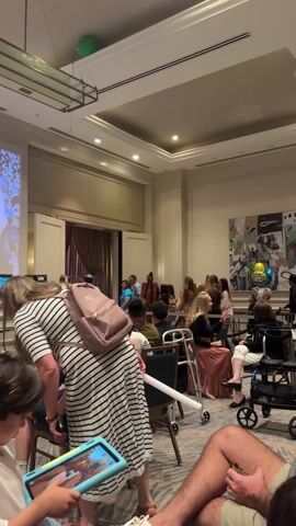 The video shows a room with a crowd of people attending a presentation. There is a screen showing a video on the left wall, with a projection device on a table. Attendees are sitting in chairs, looking at screens of their phones and tablets. Some people are sitting in walkers and wheelchairs. A woman with a striped dress and pink backpack is tending to a child seated with a tablet. A man wearing shorts is also sitting. In the background is a large painting on the wall, and a set of open doors. A green balloon floats from the ceiling.