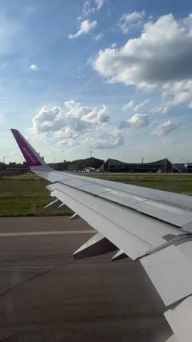 This video shows a view from an airplane window as it taxis down a runway. The wing of the plane is visible in the foreground, with a pink logo on the tail fin. In the background, an airport terminal building with a distinctive curved roof is visible, along with a control tower and grassy fields. The sky is blue with scattered white clouds. The plane appears to be accelerating down the runway.