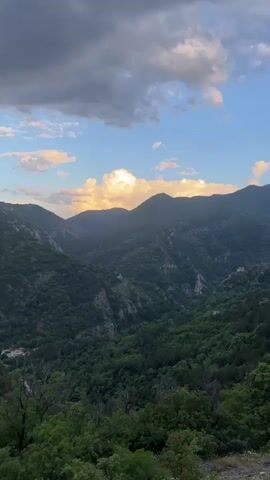 The video begins with a wide shot of a mountainous landscape under a sky dotted with clouds, some illuminated by the setting sun. The mountains are densely covered in lush green forest. The camera slowly zooms in towards a small, weathered stone building with a red-tiled roof, perched on a rocky outcrop amidst the trees.