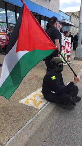 The video shows a person seated on the edge of a sidewalk, holding a Palestinian flag on a stick. The individual is wearing dark clothing, a hood, and a patterned scarf covering their face. They appear to be looking at a device in their lap. Behind them, there is a group of people, some holding signs. The setting is an urban environment, with buildings visible in the background. The sky is overcast.