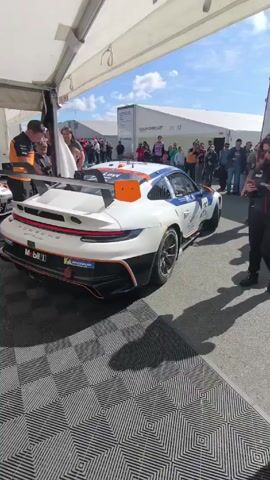 The video shows a white, orange, and blue Porsche race car being pushed out of a tent by two men wearing black pants and orange shirts with black sleeves. A woman in a black jacket is standing to the right, looking at her phone. Other people are visible in the background, beyond the tent. The car has a large wing and the logos of Mobil 1 and Michelin. The ground is covered in black and grey patterned interlocking tiles. There is a trailer behind the crowd. A black rolling box is pushed to the left and another race car is visible further down the tent.
