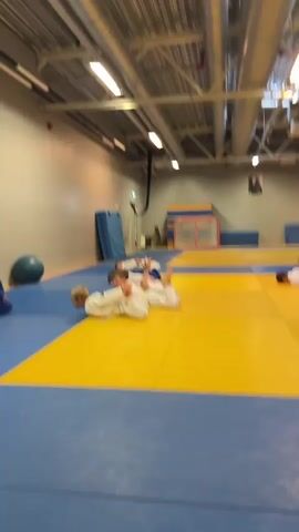 The video shows four young children wearing white judo gis on a blue and yellow mat. They are lying on their stomachs with their legs bent at the knees and their feet pointing upwards. They appear to be performing some kind of exercise or drill. In the background, there are mats leaning against a wall, a hockey net, and industrial lighting.