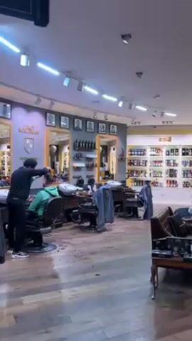 The video shows the interior of a barbershop and retail store with a sophisticated ambiance. A barber is actively cutting a customer's hair in a traditional barber chair.  The store is well-lit by both overhead fluorescent lighting and a chandelier, and features hardwood flooring. Display shelves stocked with products are arranged along the walls, and comfortable seating areas are furnished with leather couches, armchairs, and ornate tables. Decorations include portraits, mirrors with gold frames, and decorative rugs. The overall impression is one of luxury and traditional style.