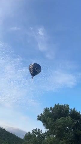 A black and white checkered hot air balloon with the word "Dentures" written vertically in white text ascends into a bright blue sky. Thin, wispy white clouds streak across the sky. The bottom portion of the balloon is visible, with a basket hanging below. The balloon is in focus and centered in the frame, while the sky fills the majority of the video. In the lower right corner, the top of a dense green tree is visible, and a hint of a distant green hill is seen in the lower left. The video appears to be a close-up shot of the hot air balloon as it travels upwards.