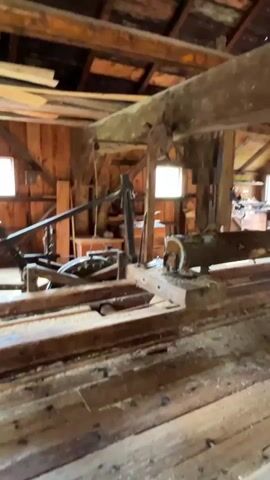 A wooden sawmill with a saw blade cutting a log is shown. The log is being fed into the saw. The sawmill is made of wood and appears to be old. There is sawdust on the floor. There are windows in the background.