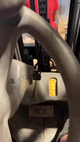 The video shows the interior of a forklift from the perspective of the operator. The main subjects are the steering wheel, dashboard, and part of the forklift's mast. A person wearing a red t-shirt is visible in the background, seated and facing away from the camera. The setting appears to be a warehouse or industrial space, with shelves and pallets visible in the background, along with some exposed brickwork.

The activity taking place seems to be the forklift being operated, as indicated by the flickering lights on the dashboard and the subtle movements of the forklift's mast. The time of day is not specified, and the weather is not discernible from the interior shot. The overall mood is functional and work-oriented.

Notable details include the prominent yellow lever on the dashboard, which likely controls hydraulic functions, and the series of indicator lights on the right side of the dashboard, one of which appears to be lit green. The visible text on the dashboard is "CROWN," indicating the manufacturer of the forklift. The close-up shots of the forklift's controls and mast suggest a focus on the machinery itself and its operation.