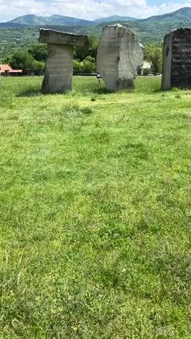The video shows a stone monument in a green grassy field. The monument has several large standing stones, some of which have writing on them. There is a horizontal stone laid across the top of two vertical stones, creating a doorway. The sky is blue with white clouds. In the background, there are green trees and mountains. A person walks near the stones near the end of the video.