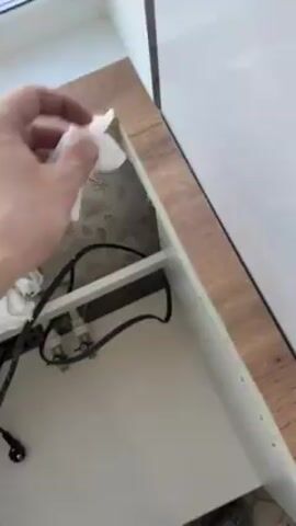 A hand holding a white tissue is wiping a wooden kitchen counter. The counter is brown with a wood grain pattern. The inside of the counter is white. Black wires and pipes are visible inside the cabinet. The base of the cabinet is white. The counter is next to a white wall. The tissue is held in the right hand of a person wearing Crocs. The person is cleaning the edge of the counter. Wallpaper with a floral design is inside the cabinet.