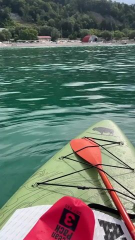 The video is shot from the perspective of someone on a paddleboard, looking forward. The paddleboard is green and has a red paddle resting on it. A red dry bag is also visible. The water is calm and a greenish-blue color. In the distance, there is a beach lined with white lounge chairs and umbrellas. Behind the beach, there are trees and hills. The sky is overcast with clouds. The scene is a coastal landscape.
