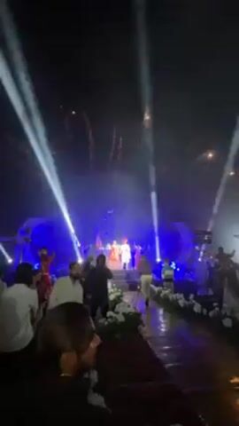 A wedding celebration unfolds on an outdoor stage, illuminated by spotlights and blue lighting. The bride, resplendent in a red and gold lehenga, stands hand-in-hand with the groom, who is dressed in a white sherwani and a red turban. Dancers in red outfits perform on either side of the couple. Observers stand in front of the stage, some recording the event with their phones. The area surrounding the stage is decorated with white floral arrangements. Fireworks are seen in the sky.
