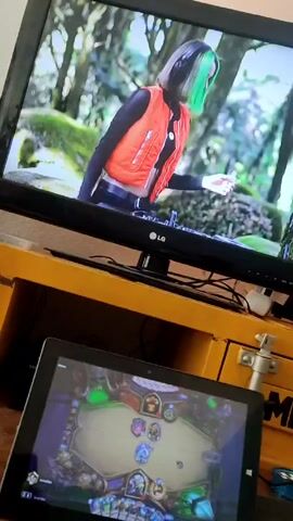 The camera faces a room with a television and a tablet. The TV is on, and the screen shows a woman with short dark hair and a streak of green standing in what appears to be a forest setting. She is wearing a bright orange vest over a black top and appears to be mixing music. Under the TV is a yellow desk-like surface. The tablet displays a card game that appears to be Hearthstone. There are game cards on the screen, and it looks like a game is in progress. A mouse is on the desk next to the tablet.