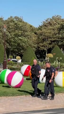 The video shows a sunny day with a clear blue sky. In the foreground, three police officers in dark blue uniforms with vests walk along a paved walkway. They are moving from left to right across the frame. Behind them, on a patch of grass, are several large, inflatable beach balls in various colors, including green, white, pink, and red. To the left of the officers, there is a lamppost with a French flag attached. Further back, there is a decorative garden bed with flowers and manicured bushes. In the background, a large, ornate red building with a grey roof stands under the trees. The building appears to be a historic structure, possibly a town hall or a chateau. The overall scene suggests a public space, perhaps a park or a plaza, decorated for an event or festival.