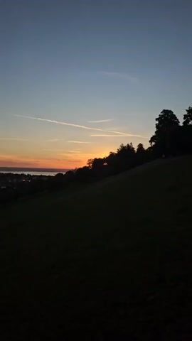A vertical video of a sunset over a rural landscape. The sky transitions from deep blue at the top to orange and pink near the horizon. Silhouetted trees and a hill dominate the foreground, with a distant town or village visible on the left. Thin white contrails streak across the sky.