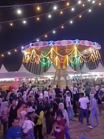 The video depicts a nighttime fair or carnival. A prominent swing ride, decorated with colorful lights and ornate trim, is visible in the center of the frame. The ride is suspended by chains and appears to be stationary at this moment. In the background, several large white tents are set up, suggesting vendors or other attractions. A densely packed crowd of people, dressed in a variety of casual attire and hijabs, are gathered around the ride and the general fairground. String lights are strung across the scene, illuminating the area and adding to the festive atmosphere. To the right of the swing ride, a tall, metal tower structure, also adorned with lights, rises into the dark sky. The ground appears to be uneven dirt or sand. The overall impression is one of a lively and bustling outdoor event.
