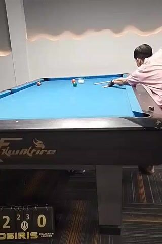 A person plays pool on a blue table. The person is wearing a pink shirt and is positioned behind the table, about to strike the cue ball with a pool cue. Several colored pool balls are scattered across the table. In the foreground, a scoreboard shows the numbers 2, 3, and 0. The environment appears to be a dimly lit room with decorative lighting on the ceiling.
