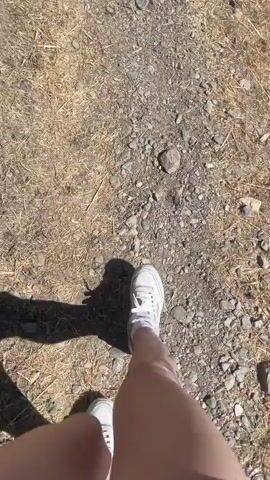 The video shows a first-person perspective of someone walking on a narrow dirt path. The path is lined with dry grass and rocks. In the background, there is a steep, rocky incline and trees. The person is wearing white sneakers and their legs are visible. The lighting suggests it is daytime and sunny.