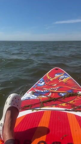 The video shows a first-person perspective of someone sitting on a paddleboard in the ocean. The paddleboard has a red and orange design with a dragon graphic on the front. The person is wearing a white Croc on their left foot, and dark leggings or swim pants. They are looking out at the ocean, which is choppy with moderate waves under a clear blue sky. Some seaweed is visible on the paddleboard. The overall scene suggests a recreational activity on a sunny day.