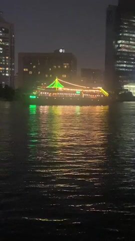 A boat is illuminated with green and yellow lights as it travels on a dark body of water. The boat has a peaked roof with lights strung along its edges. The water reflects the lights from the boat and the surrounding buildings, which are tall and modern. The sky is dark, indicating that it is nighttime. There are no people visible on the boat or in the surrounding area. The overall scene is one of urban nightlife and scenic beauty.