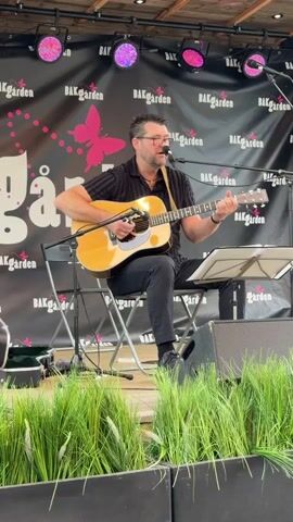 A man with glasses is sitting on a chair and playing an acoustic guitar. He is wearing a black shirt with a striped pattern and black pants. He is also wearing a necklace and a watch. The man is singing into a microphone that is attached to a stand. Behind him, there is a banner with the words "BÄKGÅRDEN" and a butterfly logo. There are also some pink lights on the ceiling. In front of the stage, there are some green plants. The man is playing music on a stage.