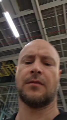 A bald, bearded man wearing a black collared shirt is in an indoor industrial or warehouse setting. The ceiling has visible fluorescent lighting and exposed beams. In the background, there is metal shelving and some yellow and green infrastructure. The man is looking directly at the camera. He makes a peace sign with his right hand at 00:01 and then lowers it. He speaks briefly at 00:02.