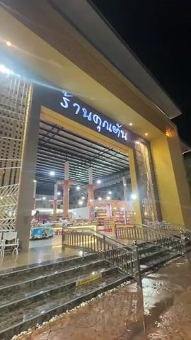 The video shows the exterior of a large building at night, likely a tourist stop or rest area. The building has a large entrance with a sign in Thai that says “Raan Khun Tan.” The area in front of the building has stairs leading up to the entrance, along with an ice cream stand featuring pink neon lights. Several tour buses are parked nearby. People can be seen walking around the area. The scene appears to be well-lit.
