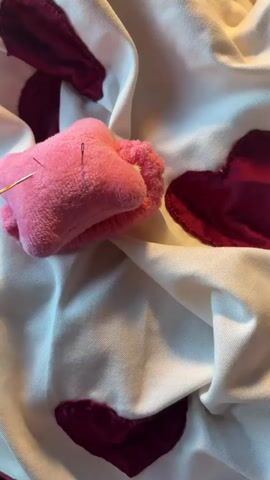 The video shows a close-up of a person's hand, with manicured nails, interacting with a pink star-shaped object that appears to be a pincushion with needles. The pincushion is resting on a white fabric that is decorated with several dark red heart shapes. The hand is moving and adjusting the pincushion, with fingers delicately touching it and the needles. The background is softly blurred, but the texture of the fabric and the sheen of the hearts are visible. The lighting suggests an indoor setting. The action is focused on the small object and the hand's precise movements.