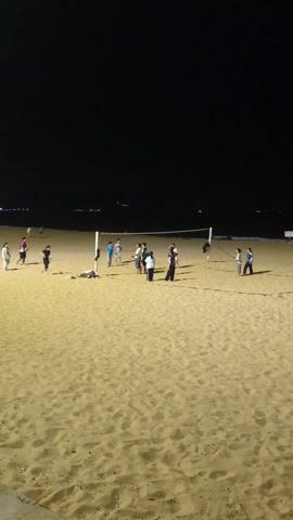 At night, a group of approximately a dozen people are gathered on a sandy beach playing volleyball. A net stands in the middle of the group.  The beach is well-lit, and the figures are silhouetted against the dark sky.  In the distance, faint lights suggest a city skyline. Some people stand still, while others are actively moving around the net, presumably during play.