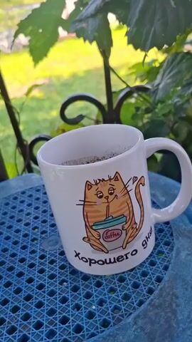 A white mug with an orange cartoon cat holding a cup of coffee sits on a blue plastic table. The cat has closed eyes and a heart above its tail. The mug has text below the cat that appears to be in Russian. The background is blurred, showing green leaves and sunlight. The mug is slightly tilted.
