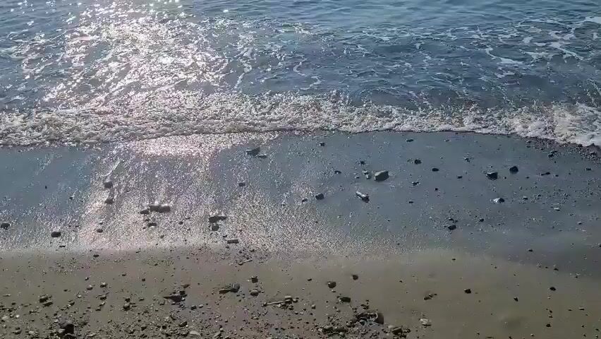 The video shows a view of the ocean and a sandy beach from a slightly elevated perspective. Gentle waves are lapping at the shore, with sunlight sparkling on the water's surface. The beach appears to be made of sand and small pebbles. There are no people or animals visible in the video. The overall scene is peaceful and natural.