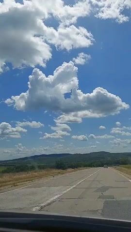 The video shows a view from inside a vehicle traveling on a paved road. The sky is blue with fluffy white clouds, and a large cumulus cloud formation is prominent in the center. The road stretches ahead, with a white line dividing the lanes. The surrounding landscape consists of rolling hills covered with green trees and dry, grassy fields. The sun appears to be shining brightly. A white car is visible in the distance on the road. The bottom of the video shows a dark dashboard or interior of the vehicle.