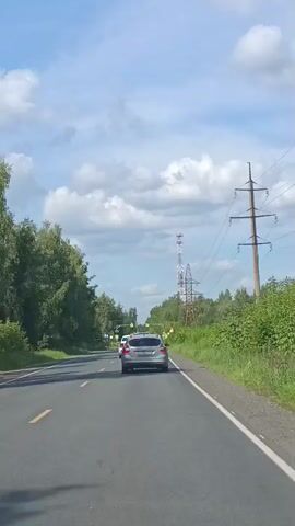 A silver car is traveling down a two-lane road with a tree line and power lines on either side. The sky is blue with fluffy white clouds. The camera is mounted inside a vehicle, looking out through the windshield. The road is paved with asphalt and has a yellow line marking the center. There are other cars on the road, including a dark-colored bus and a white sedan ahead of the silver car. The silver car appears to be traveling at a moderate speed.