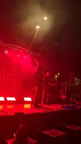 The video shows a dimly lit outdoor concert stage at night. Red stage lights cast a dramatic glow, with some fog or smoke present. Two musicians are visible on stage. The primary focus is on a lead singer or frontman, standing center stage, holding a microphone. They are wearing dark clothing, possibly with patterned pants. To their right and slightly behind, another musician appears to be playing a guitar. A drum set and at least one other guitar are visible on stage. The background is dark, with faint outlines of trees and buildings. The foreground shows the edge of the stage with some equipment, possibly speakers or amplifiers. The overall atmosphere is that of a live musical performance.