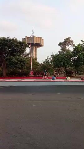The video opens on a wide shot of a street with a tall water tower in the background. Several trees and a red and white striped pole are visible in the middle ground. Two people are walking on a sidewalk, one of them pushing a cart of colorful objects. A light blue truck drives past, followed by a scooter with two riders. The video then pans to show more traffic, including cars and a motorcycle. The scene appears to be a public area with some greenery and a noticeable water tower.