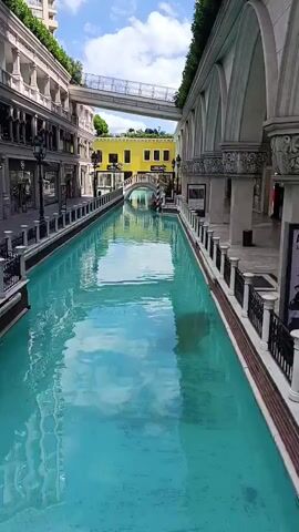A still video displays an outdoor view of a canal with turquoise water, flanked by buildings with white facades and arched windows. On the left side, a row of shops with display windows lines the canal. On the right side, a covered walkway with arched openings is visible. A white arched bridge spans the canal in the background, leading to a yellow building. A modern glass-enclosed pedestrian bridge stretches across the scene above the canal and buildings. The sky is blue with scattered white clouds, and green foliage is visible on top of some buildings. The water reflects the sky and buildings. There are no people clearly visible in the foreground.
