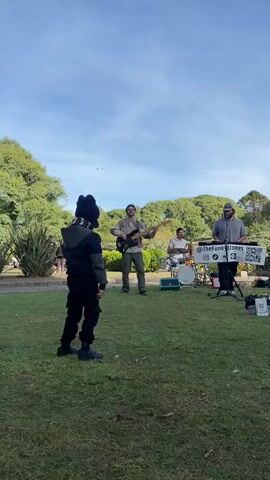 Four men perform as a band on a grassy area in front of a group of onlookers. The band has drums, a keyboard, and a bass guitar. The group's name, “The Funk Stones,” is visible on a sign. One woman dances in front of the band. A child in a winter hat watches the band and the dancing woman. Trees and green foliage can be seen in the background. The sky is blue and partly cloudy.