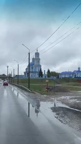 A video shows a blue and white church on a small hill next to a wet road, shot from a moving vehicle. The road is wet with puddles, and cars can be seen driving on it. The church has a tall bell tower and a blue dome, and it sits on a grassy hill surrounded by a blue fence. Power lines run parallel to the road. There are also lampposts, a guardrail, and a water pipe. The sky is overcast and gray.