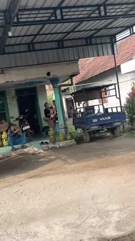 The video opens on a scene under a covered outdoor area. A blue trailer with the word "VIAR" is parked on the right side of the frame. A person is sitting on the ground to the left, wearing a blue shirt and jeans, with their legs crossed. There are several yellow propane tanks near them. In the background, a motorcycle is parked. A group of people, including one holding a baby, are standing near the entrance of a building. The building appears to be a shop or a home. The ground is dirt and gravel. The overall lighting suggests daytime.