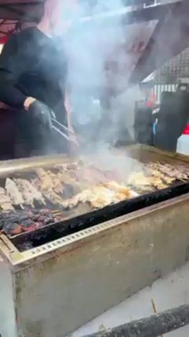 A man is seen grilling meat skewers on a large, flat grill. He's wearing a black shirt, black gloves, and sunglasses. Smoke rises from the grill as he uses tongs to turn the skewers.  He then squeezes a bottle of liquid, likely oil or marinade, onto the grilling meat. A sign reading "Daniel's BBQ" is partially visible in the background. The scene appears to be outdoors under a red canopy.