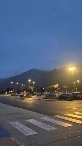 The video shows an outdoor scene at dawn or dusk, focusing on a parking lot with mountains in the background. The sky is a deep blue, suggesting either early morning or late evening. Streetlights illuminate the parking lot, casting a warm glow on the parked cars and the asphalt. Palm trees are interspersed among the streetlights. A pedestrian crosswalk is visible in the foreground, marked by white stripes. The mountains in the background are partially obscured by clouds or fog, adding to the tranquil atmosphere of the scene. The parking lot is moderately full with a variety of vehicles. The camera slowly pans from left to right.