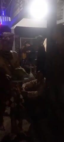 A low-light video shows a group of people gathered outdoors at night. In the foreground, a person in traditional attire, possibly a dancer or performer, is holding a plate with food items, including what appears to be a coconut. This individual is adorned with gold jewelry and a patterned skirt. The background is dark, but suggests an outdoor setting with some structures and plants. Other people are visible in the background, some standing and others sitting. There are lights in the background, possibly from a building or decorations, casting a dim glow on the scene. The overall impression is of a cultural event or gathering taking place after dark.