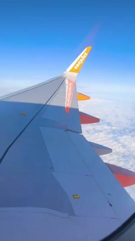 This is a first-person perspective video taken from an airplane window. The video shows the wing of the airplane, which is grey and white with orange accents and the EasyJet logo on the wingtip. Below the wing, there are white clouds and a clear blue sky. The video is static, with no visible movement other than what may be perceived as slight camera shake. There are no people visible in the video. The environment is a flight at altitude.