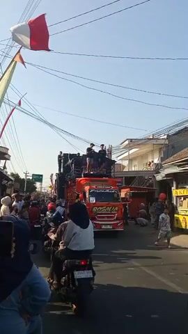 A bright, sunny day with a clear blue sky is depicted. A large orange truck, adorned with speakers and lights, is the central focus of the image. Several individuals are standing on top of the truck, appearing to be performers or sound technicians. A red and white flag, possibly the Indonesian flag, is flying from a pole to the left of the truck. The truck is moving down a street lined with buildings and power lines. A crowd of people, many on motorcycles, are gathered along the side of the street, watching the truck. The scene suggests a festive event or parade.
