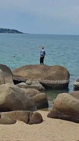 A person stands on a large rock in the ocean, holding a fishing rod. The person is wearing a hat, a patterned shirt, and dark pants. There is another person swimming in the background. The scene is a beach with sand and large rocks. The water is blue and the sky is overcast.