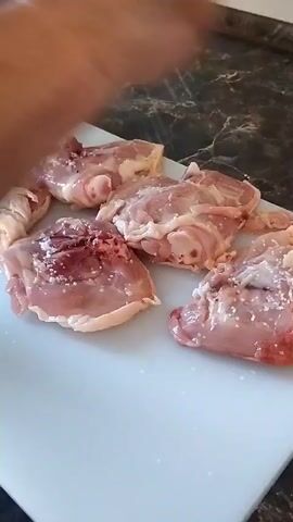A close-up shot shows several raw chicken thighs on a white cutting board. The chicken pieces are lightly seasoned with salt and pepper. A hand is visible in the upper left corner of the frame, appearing to be adding more seasoning to the chicken. The cutting board rests on a dark, speckled countertop. The lighting is bright, illuminating the scene.