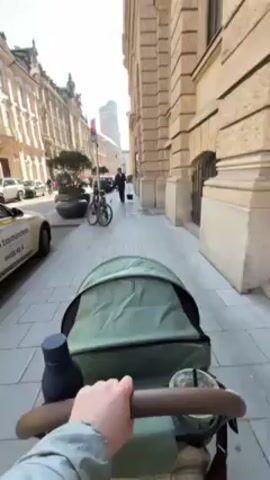 A first-person perspective video shows a person pushing a stroller on a sidewalk next to a beige stone building on a sunny day. The stroller is light green and has a dark brown handle. There is a drink and a water bottle in the stroller. Across the street, there are other buildings and parked cars. A person in dark clothing is walking away from the camera down the sidewalk. A bicycle is parked on the sidewalk. A taxi is parked on the side of the road. A landmark with two green-domed towers can be seen in the distance.