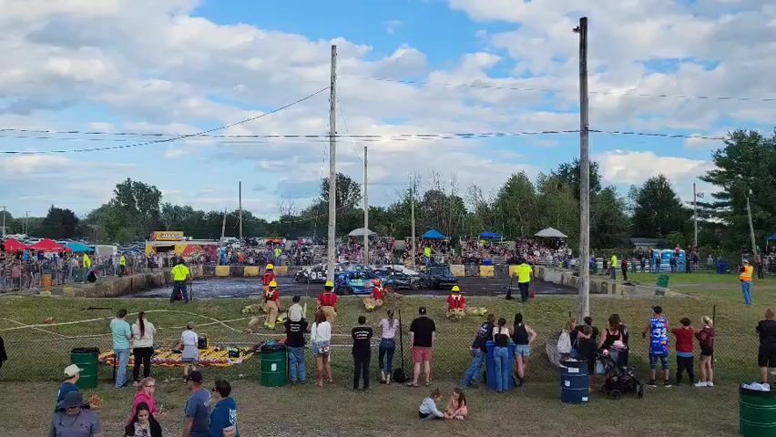 The video shows an outdoor event with a dirt track surrounded by spectators. Several vehicles, possibly for demolition derby or monster truck events, are on the track. Some individuals in safety vests and hard hats are present near the track, possibly as safety personnel or participants. A crowd of people, including children, are gathered on the grass, watching the event. Tents and other temporary structures are visible in the background, suggesting a fair or festival atmosphere. The sky is partly cloudy.